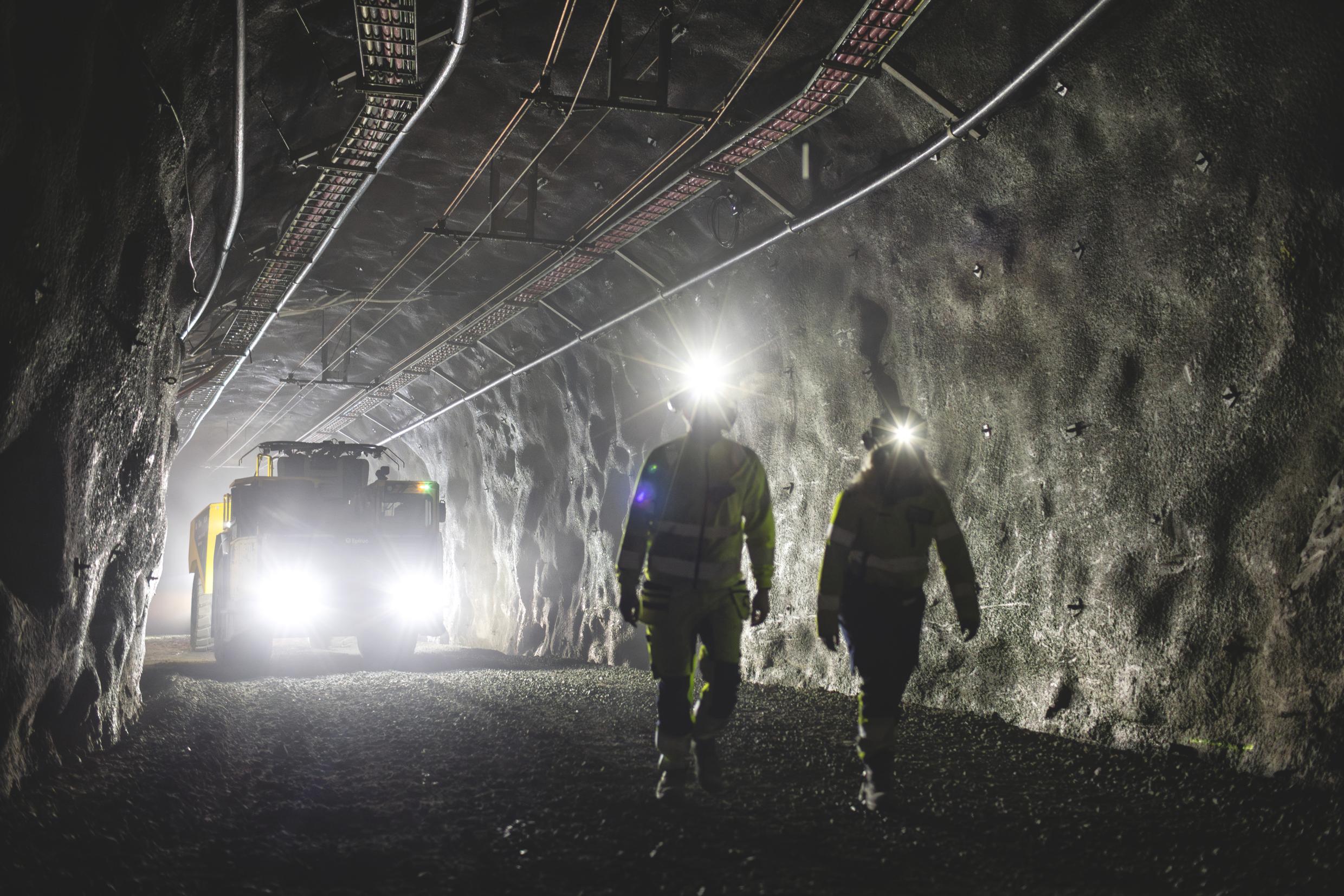 Two workers walk in front of a heavy-duty electric vehicle in an underground mine tunnel.