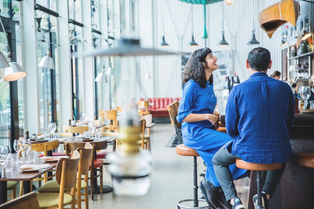 Inside a restaurant with wooden tables and chairs along a window wall. A woman and man is sitting at a bar to the right. Blurry in the front you see an oil lamp hanging from the ceiling.
