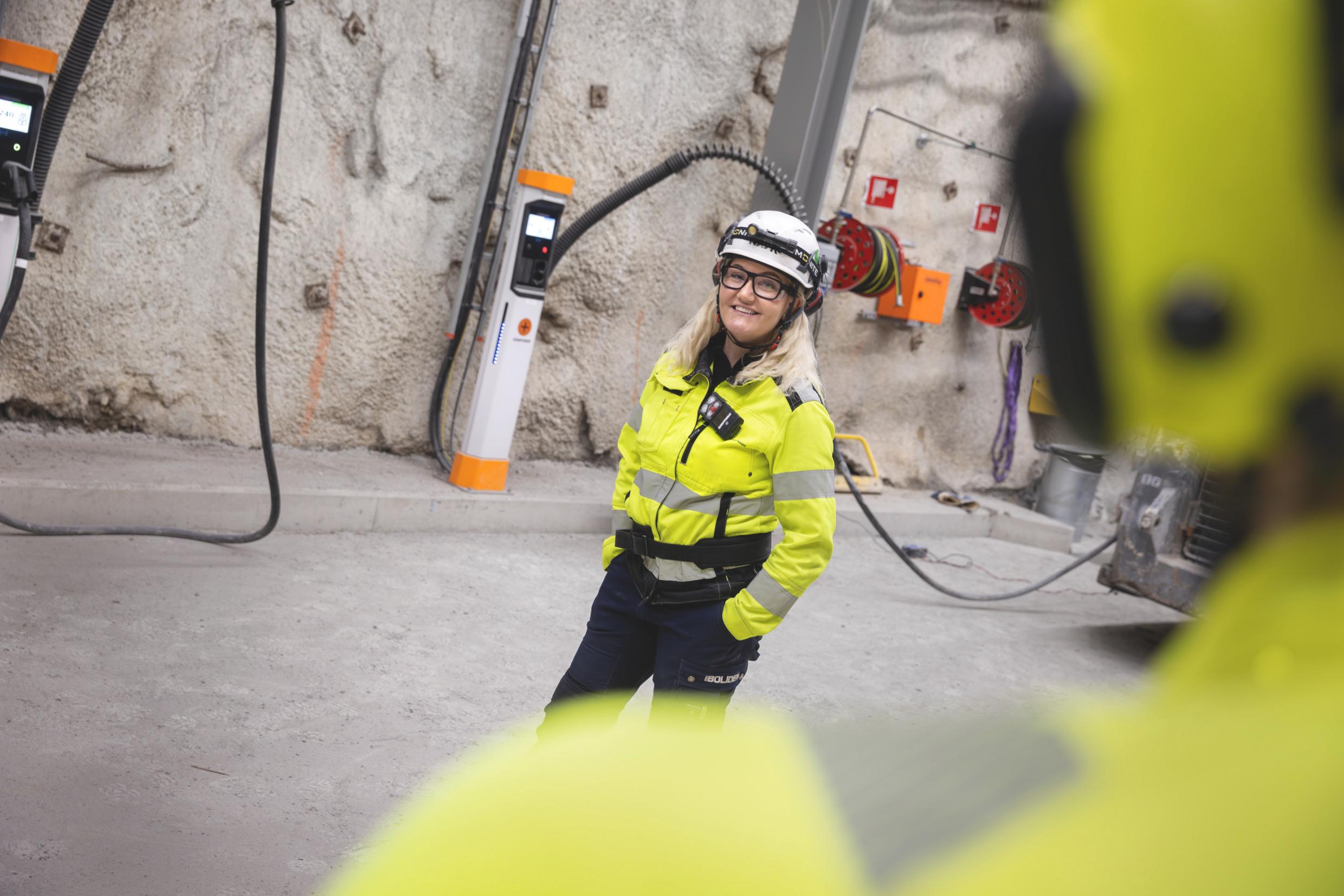 A woman wearing a high-visibility vest and a helmet stand in front of electric vehicle chargers in an underground mine.