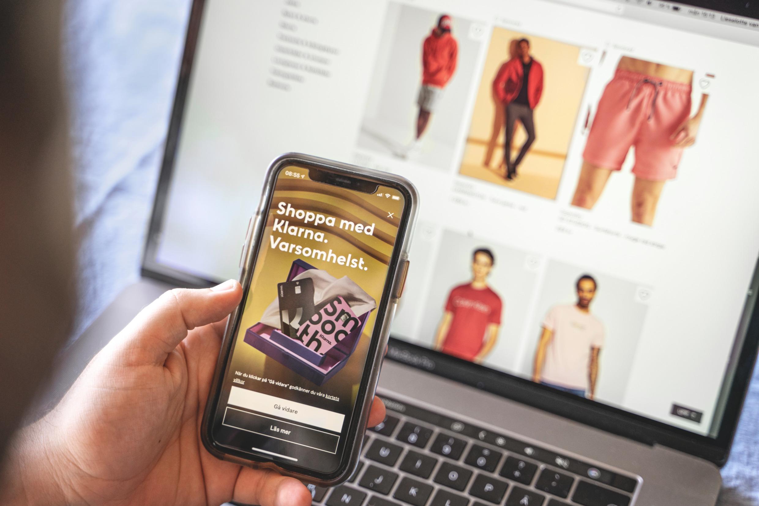 Close-up of a hand holding a mobile phone showing the Klarna app in front of a computer showing an online shopping site.