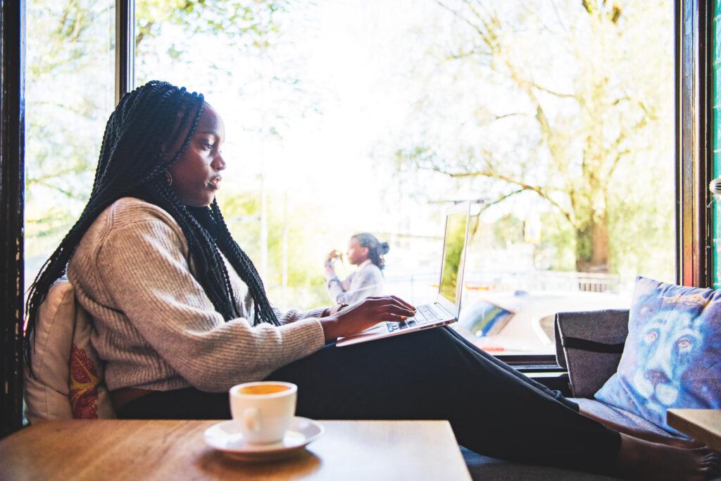 A woman is sitting inside by a window with a cup of coffee, typing on a laptop in her lap.