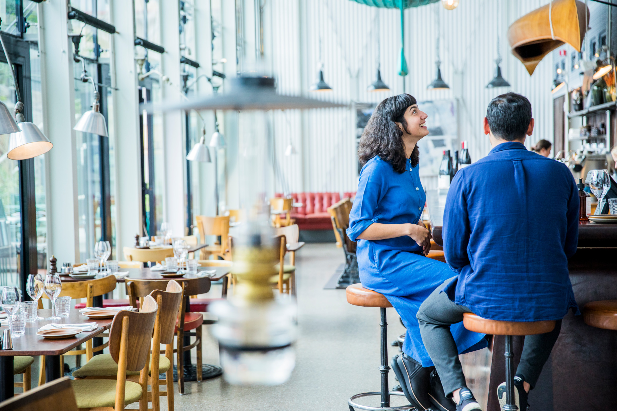 Inside a restaurant with wooden tables and chairs along a window wall. A woman and man is sitting at a bar to the right. Blurry in the front you see an oil lamp hanging from the ceiling.