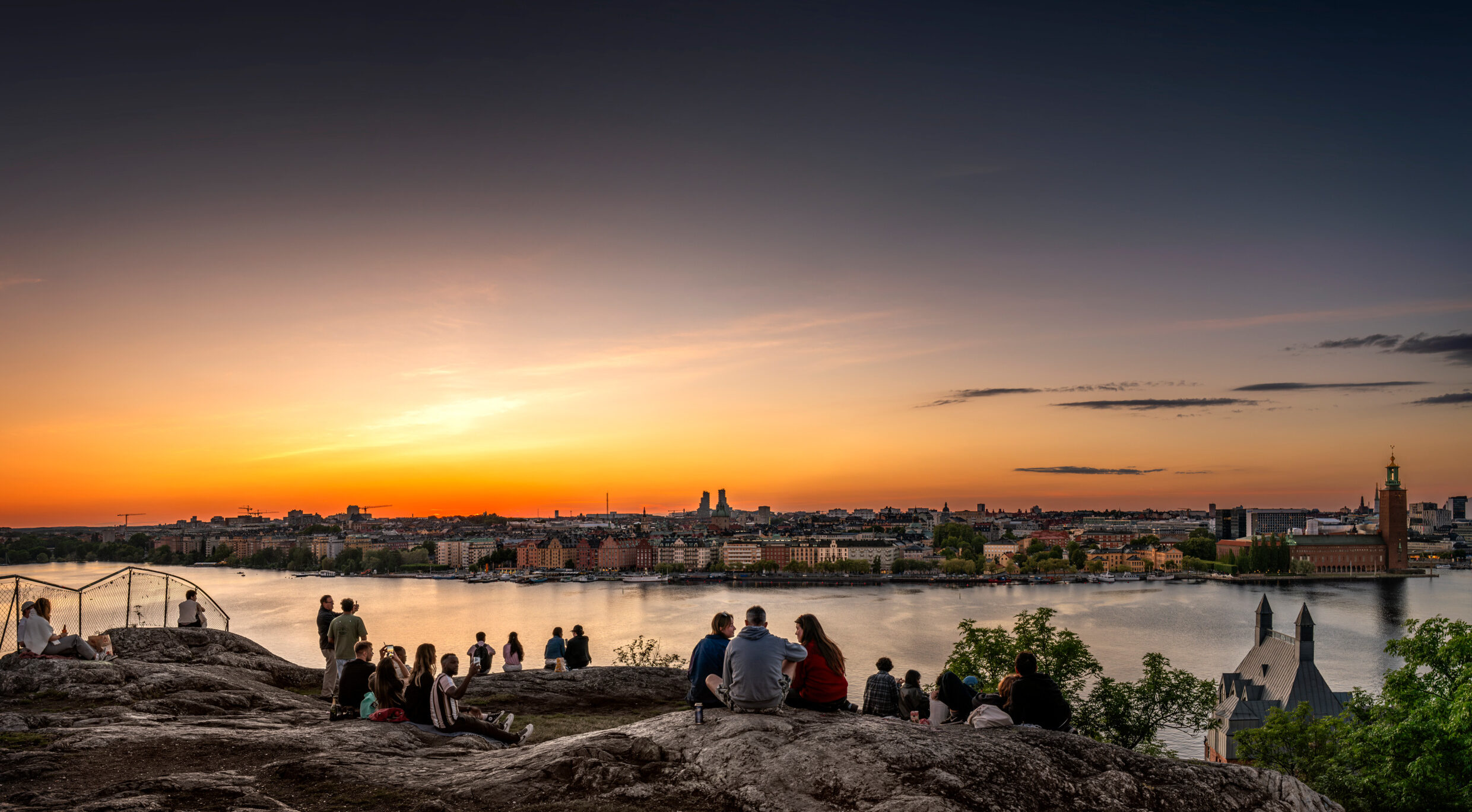 People sitting and standing on a cliff overlooking water and a city.