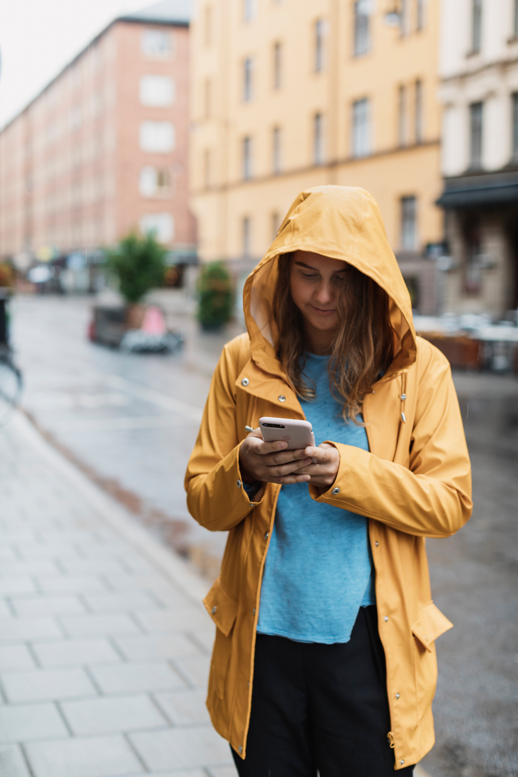 A young woman wears a raincoat and uses her phone on a city street.