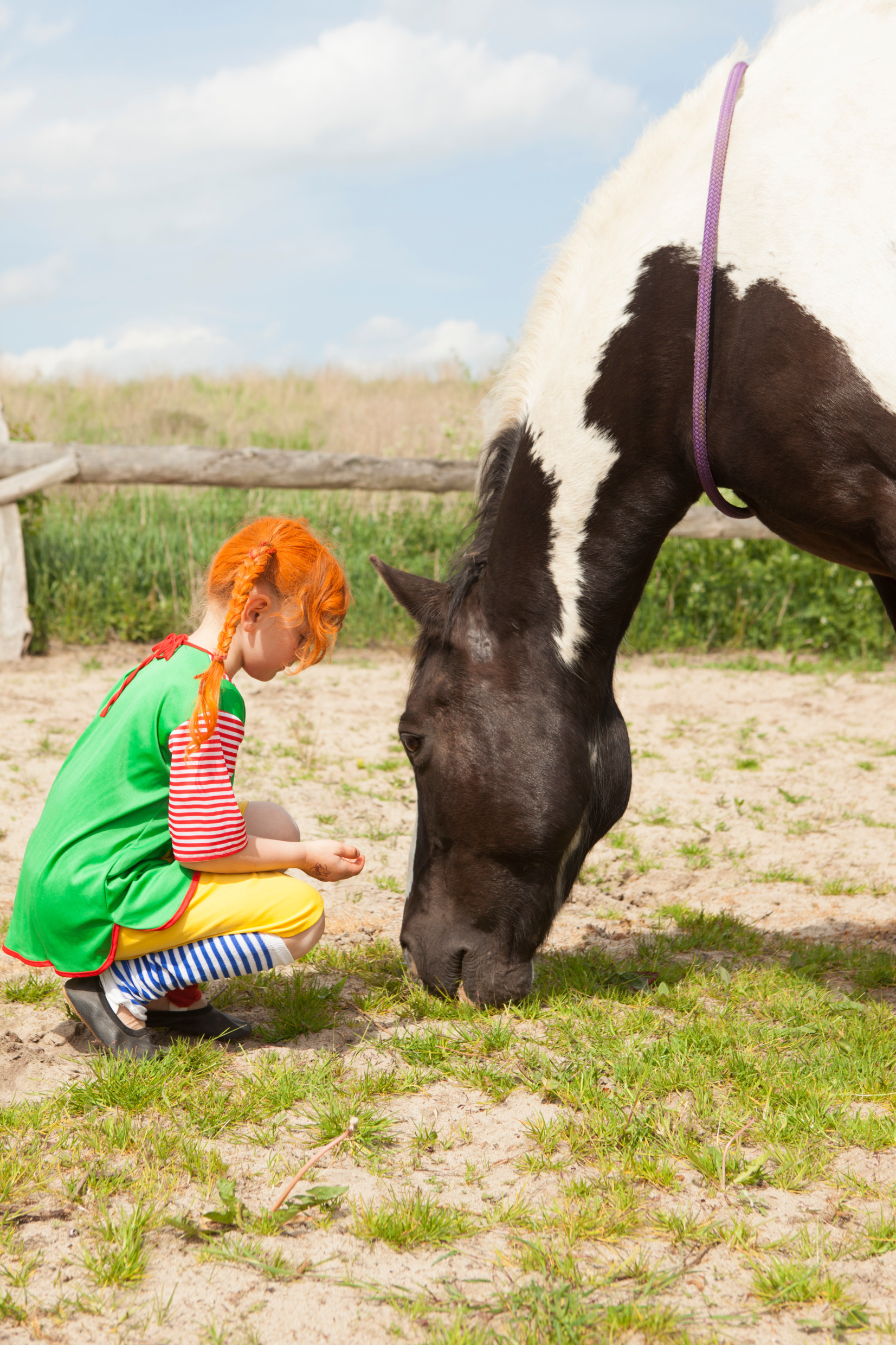 A child wearing a Pippi costume with red hair kneels in front of a horse.